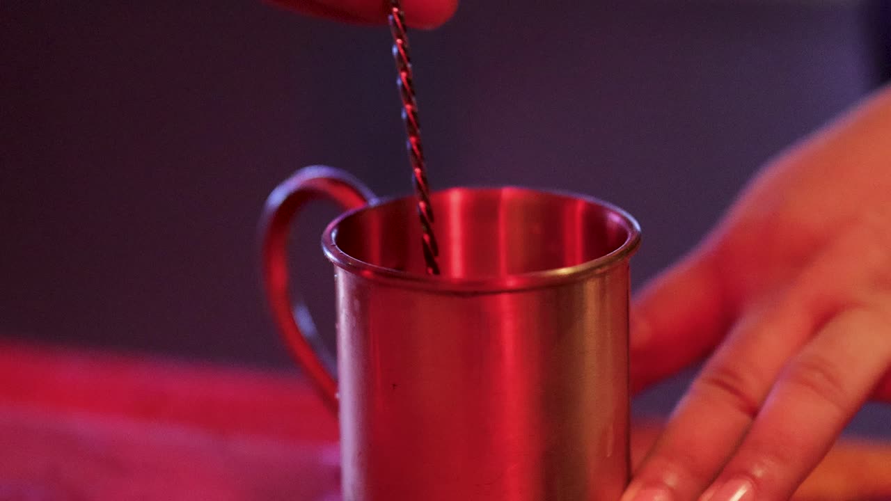 Bartender stirs drink with bar spoon in metal mug under warm, colorful bar lighting, close-up