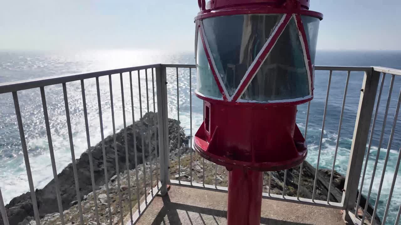 Walking down to the light inside fenced staircase and overlooking sunny ocean with waves and cliffs, turning towards cliffs and coastline and back to lighthouse keepers quarters. Ireland.