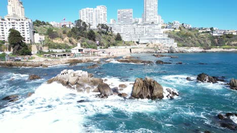 Olas-Del-Mar-Golpeando-Las-Rocas-Costeras-En-Viña-Del-Mar-Con-Edificios-Al-Fondo,-Chile