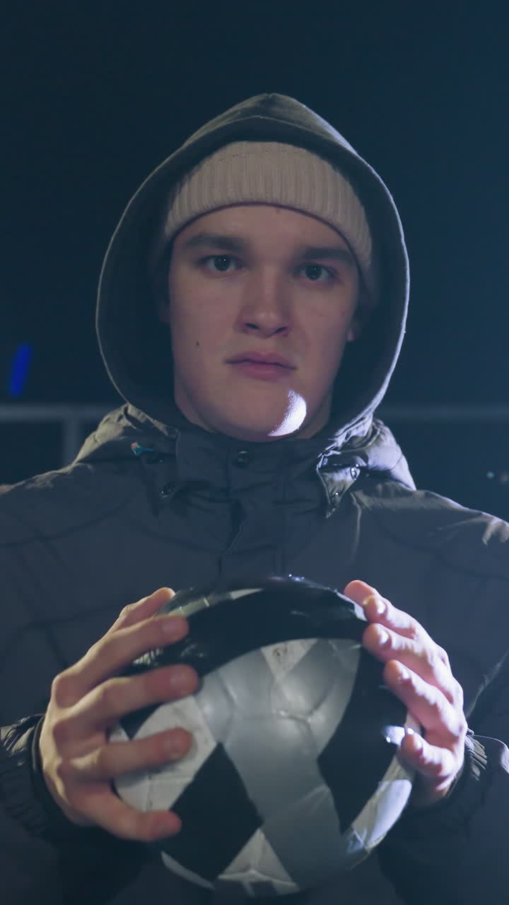 Young boy wearing hoodie holding ball, looking sad under bright streetlight during nighttime, reflecting emotional struggle and deep thought