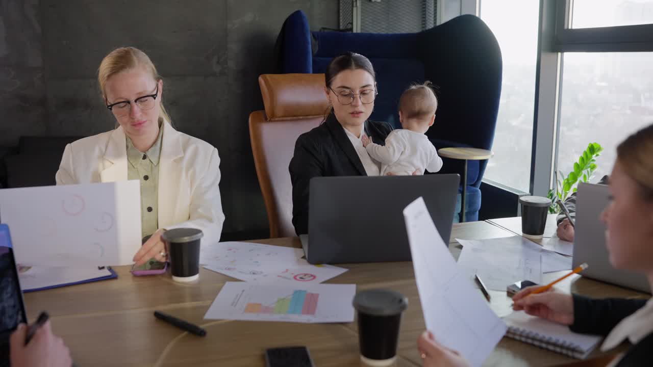 A group of businesswomen in business suits are having a meeting while a colleague is holding a small infant child in her arms while sitting at a table in front of a laptop in a modern office