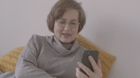 senior woman with eyeglasses sitting on bed making a video call on a smartphone 1