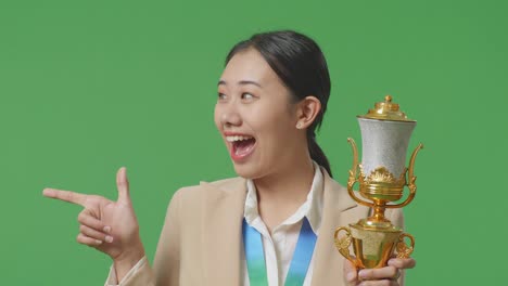 close up of asian business woman in a suit with a gold medal holding a gold trophy in her hands, smiling, and pointing to side on green screen background in the studio