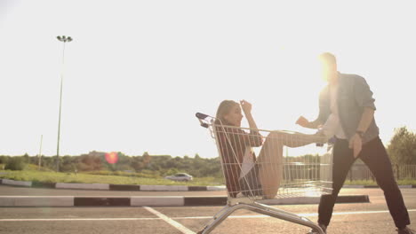 Side-view-of-a-young-man-and-woman-having-fun-outdoors-on-shopping-trolleys.-Multiethnic-young-people-racing-on-shopping-carts.-On-the-parking-zone-with-their