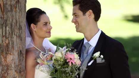 recién casados felices de pie en el parque junto a un árbol