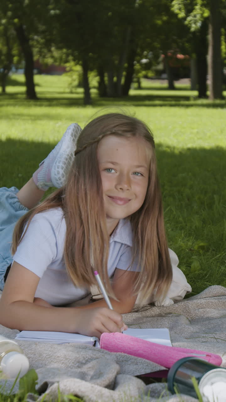 A young girl writing in a notebook while lying on the grass in a park
