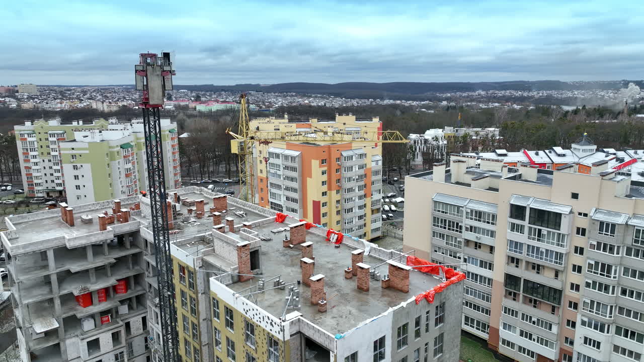 Construction cranes towering above the house in construction. Beautiful residential area full of modern apartment blocks. Cityscape at the background.