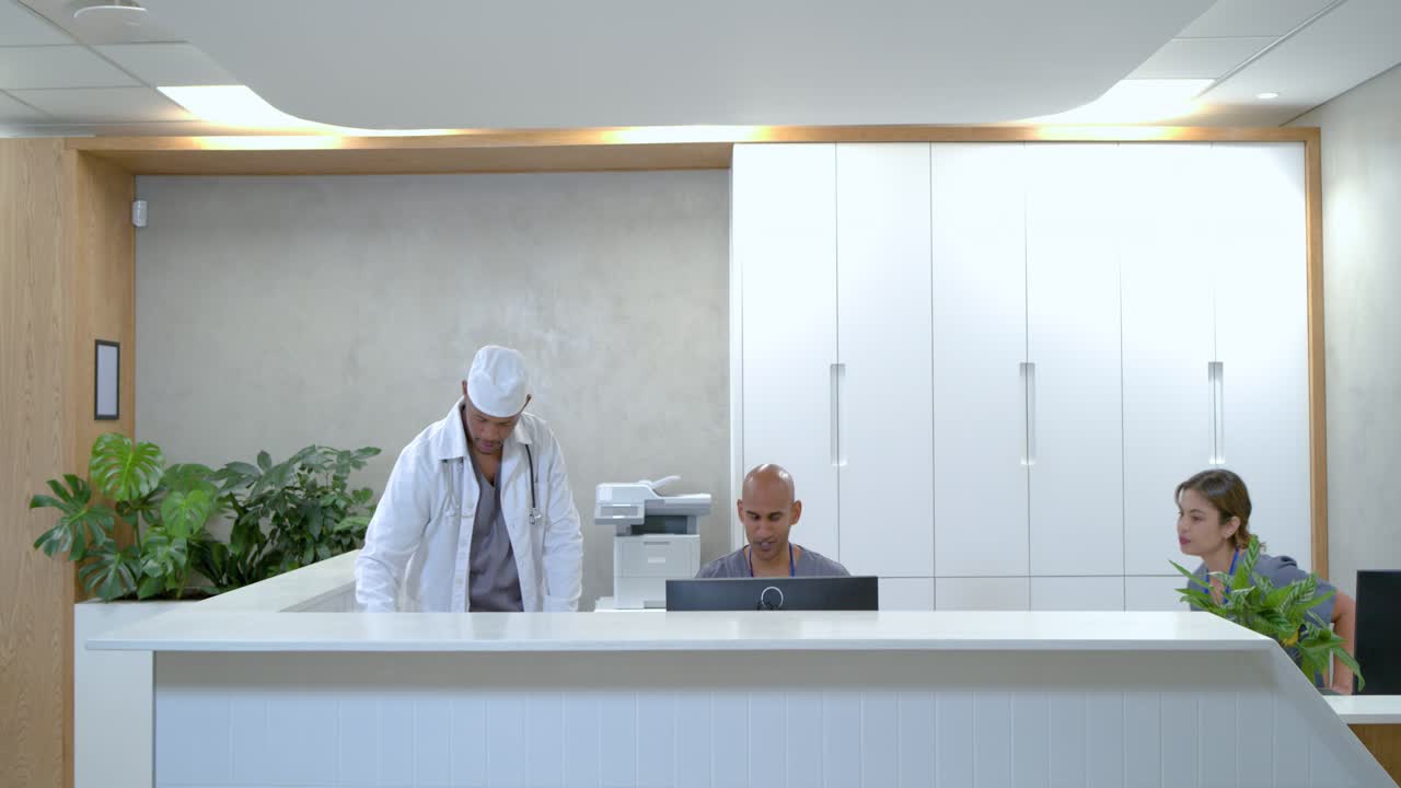 Staf resepsionis klinik yang beragam sedang berbicara di telepon, berhenti sejenak saat petugas klinis menyerahkan papan klip di konter.