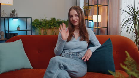 Young-woman-smiling-friendly-at-camera,-waving-hands-gesturing-hello,-hi,-greeting-at-home-on-couch