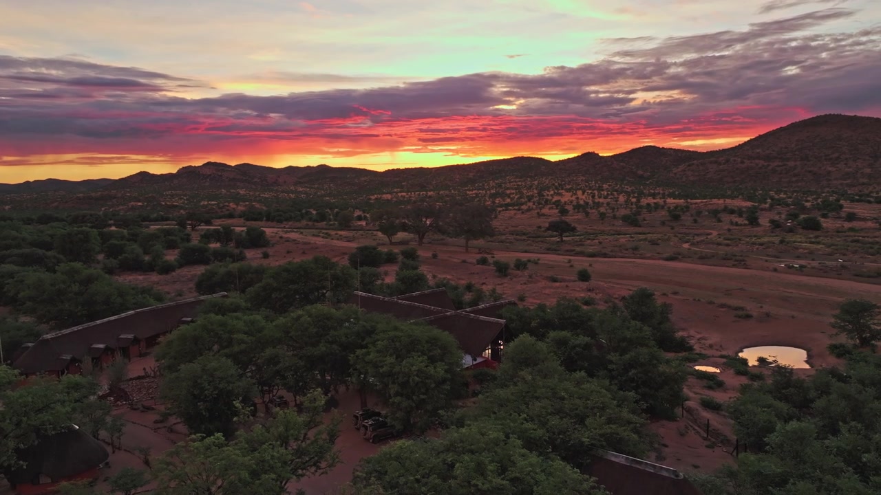 Riprese con drone di un cielo rosso incandescente su una fattoria rurale negli altipiani della Namibia, con alberi sparsi e sentieri che svaniscono nella luce della sera.