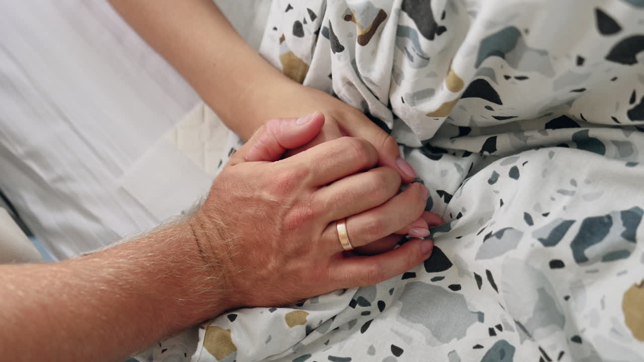 Male hand with a wedding ring covering the female one. Spouses' hands lying on the bedding. Close up.