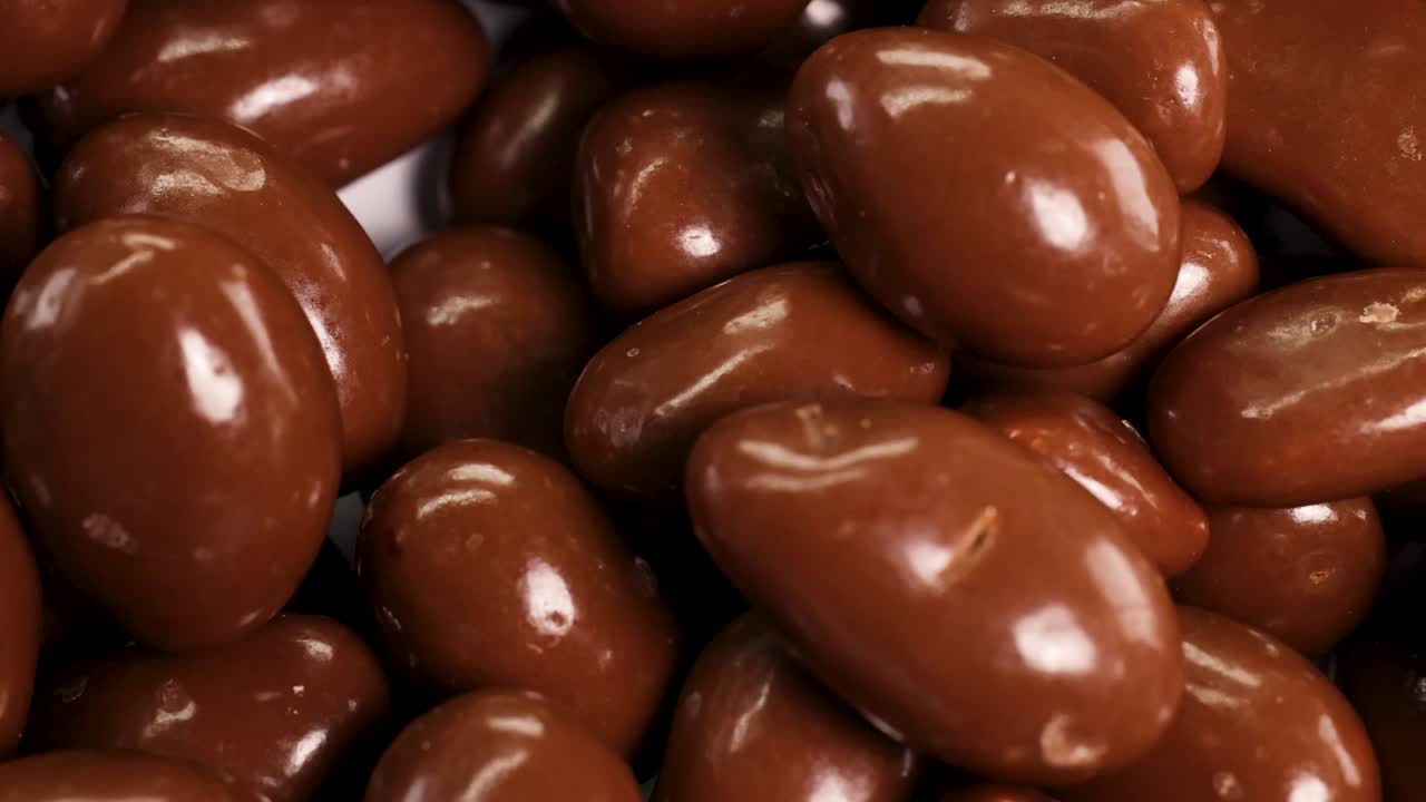 A close-up view of glossy chocolate-coated peanuts rotating under bright lighting, highlighting their smooth texture and rich color