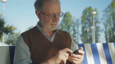 Good-looking-old-man-sitting-on-bench-outside,-typing-on-phone