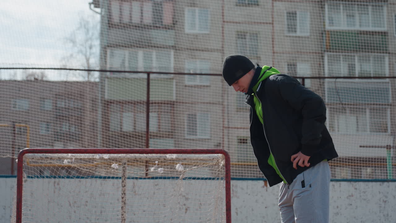 골대 옆 도시에서 생각에 잠긴 젊은이, 도시의 작은 골대 근처에서 홀로 생각하는 아이, 겨울 동안 도시의 열린 공간에 있는 작은 스포츠 골대 옆에 서 있는 남성
