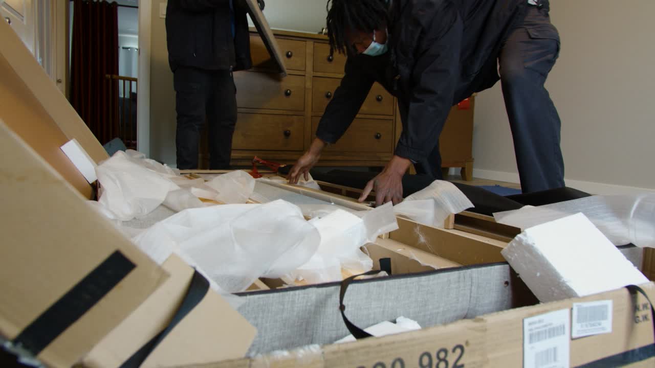 Two delivery workers unpacking large box in customer’s home; cardboard and foam pieces scattered on floor