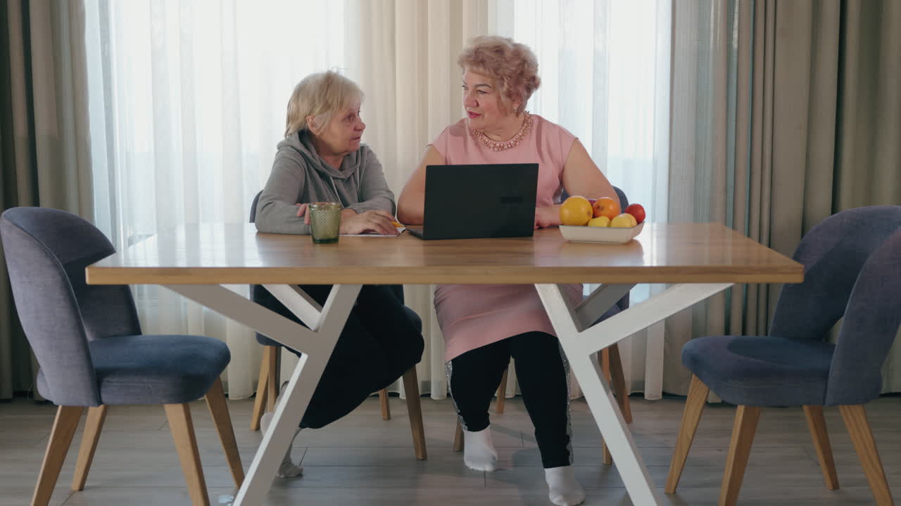 두 명의 노인 여성이 식탁에서 노트북을 사용하고 있습니다.