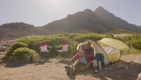 Happy-senior-biracial-couple-sitting-at-tent-in-mountains-and-embracing-on-sunny-day,-in-slow-motion