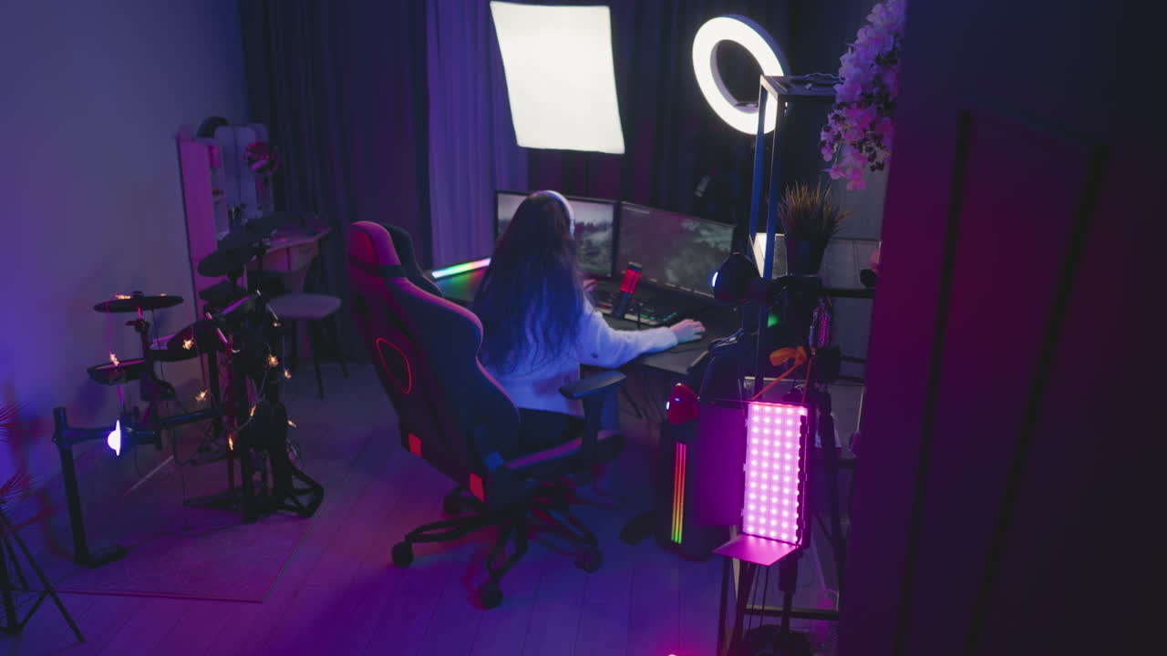 rear view of woman seated in ergonomic gaming chair wearing white headset and fuzzy sweater intensely playing video game on dual monitors using keyboard mouse beside red condenser microphone