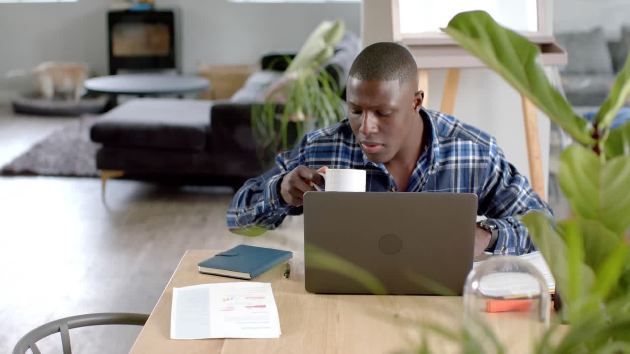 African american man sitting at table using laptop and drinking coffee at home, slow motion