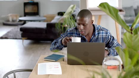 Hombre-Afroamericano-Sentado-En-La-Mesa-Usando-Una-Computadora-Portátil-Y-Tomando-Café-En-Casa,-Cámara-Lenta