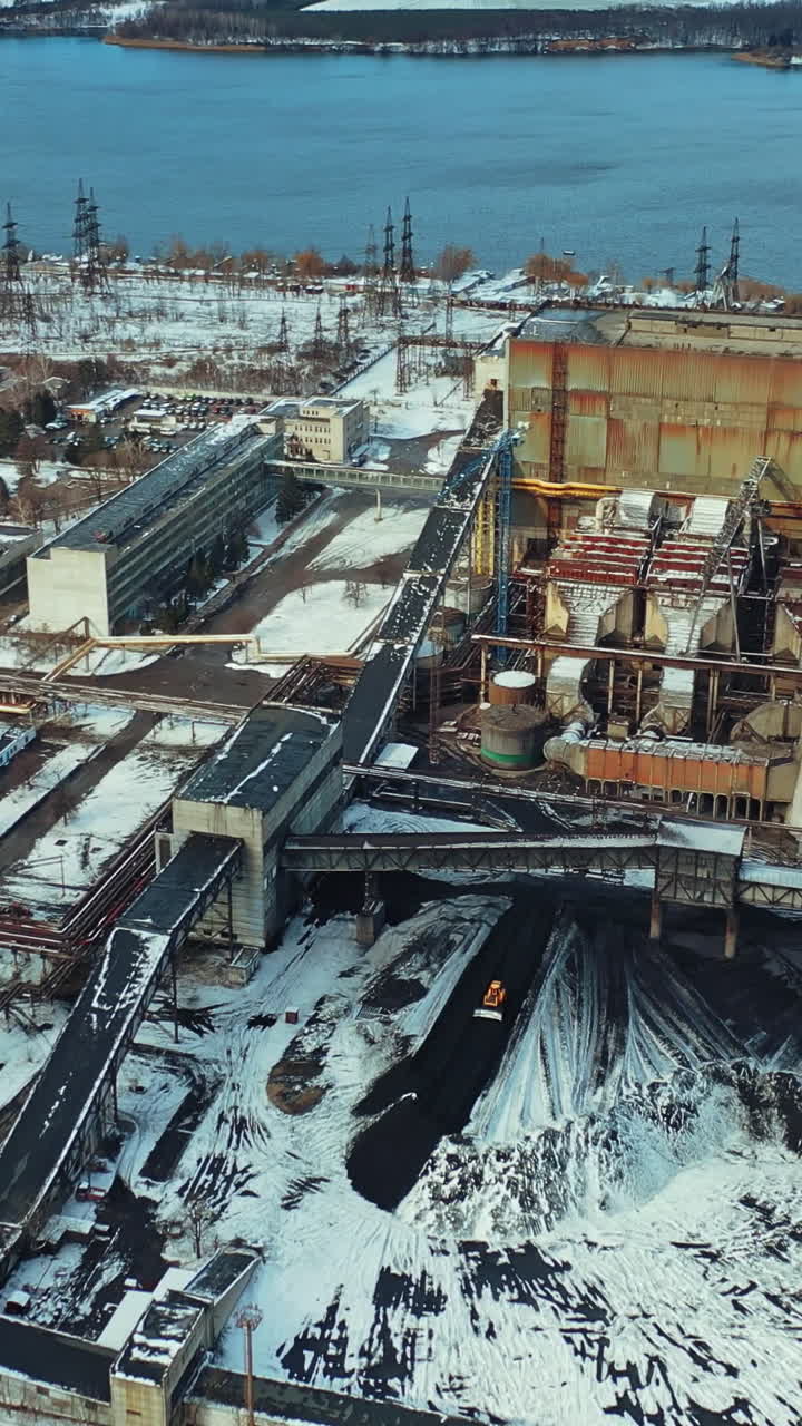 Aerial view of the vast territory of the thermal power station with administrative buildings, workshops and heaps of ash from coal, which excavator levels on the background of river behind of it. Vertical video