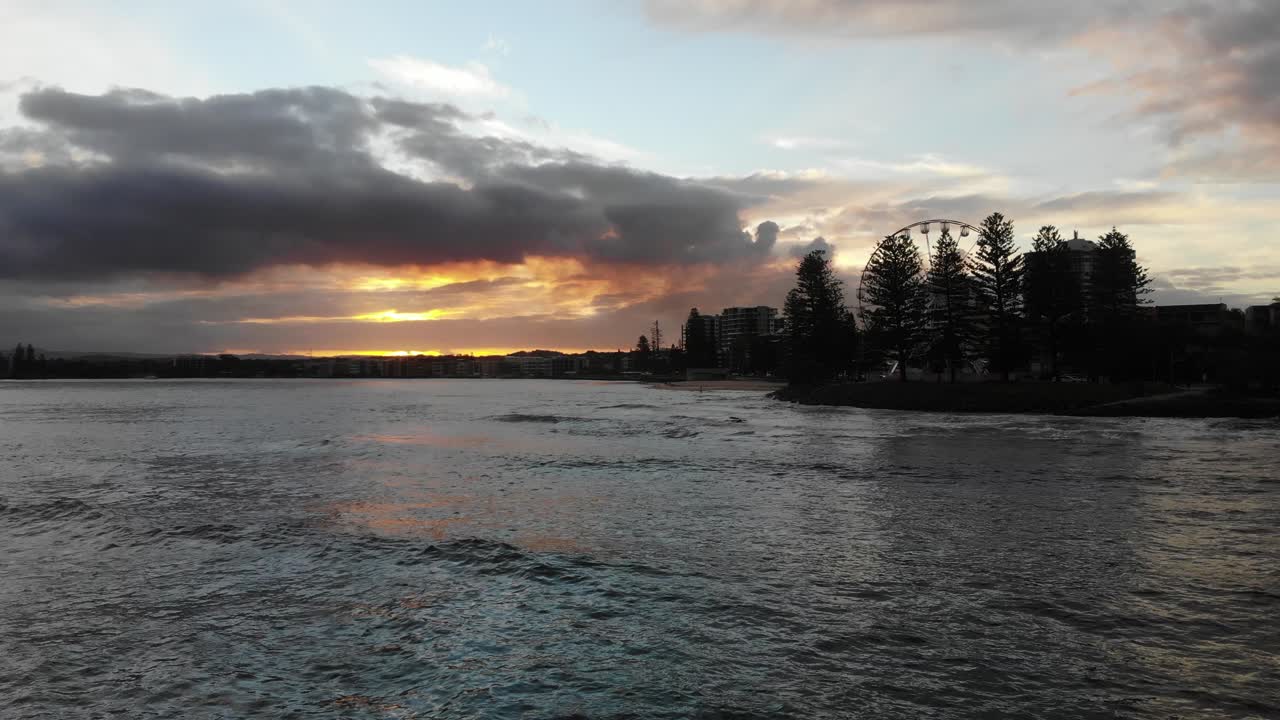 un océano en calma se sienta debajo mientras un sol cálido quema a través de las nubes en su descenso detrás de un pueblo de playa que tiene una pequeña rueda de la fortuna escondida detrás de algunos árboles de playa
