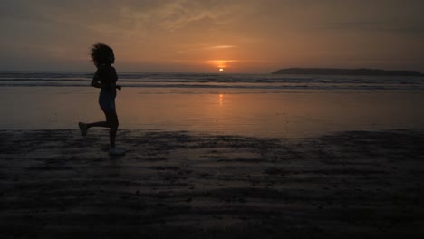 landscape view of a silhouette of a young woman running on a sandy beach by the ocean, at sunrise