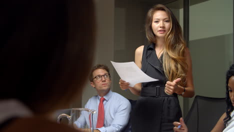 asian woman stands to address colleagues at business meeting