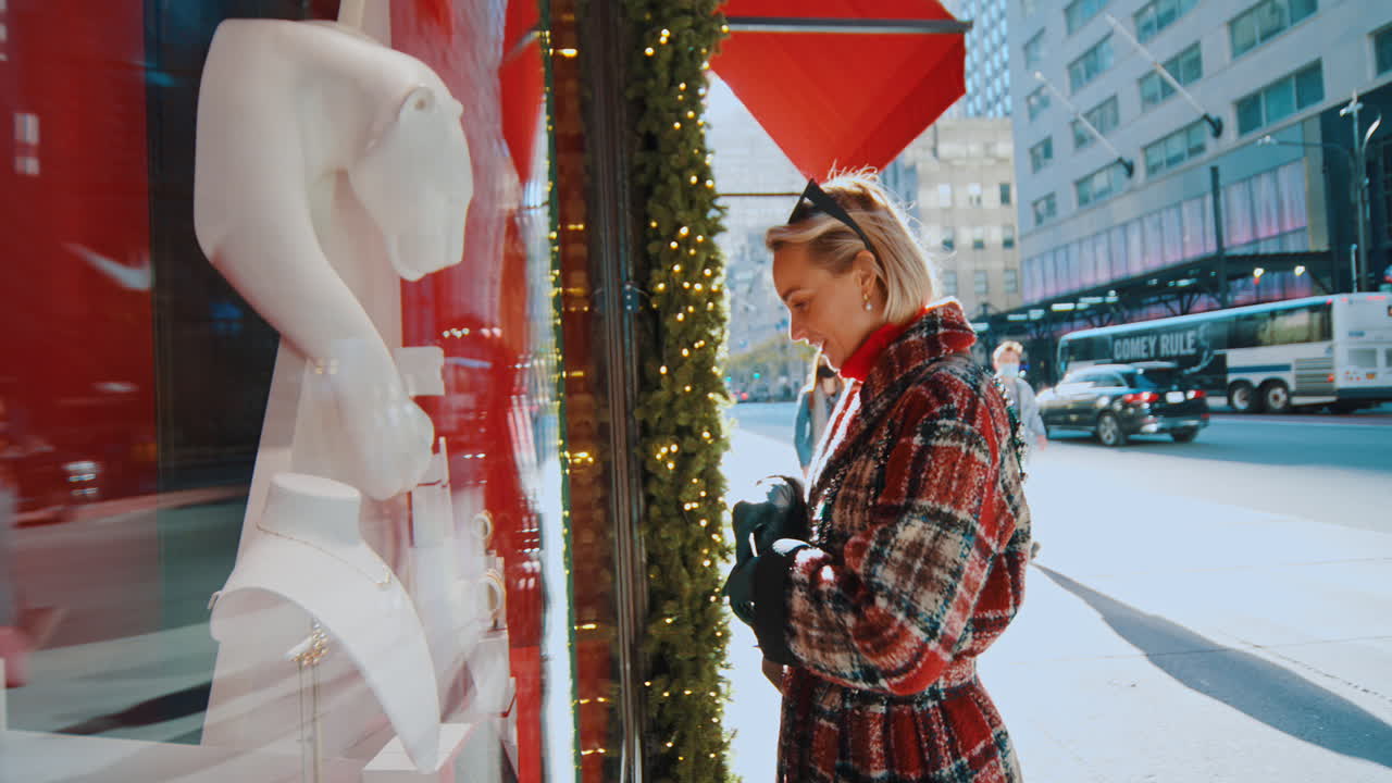 New York, USA, 5 ottobre 2025: Donna che guarda una vetrina di gioielli. Primo piano di una donna sorridente con un cappotto a quadri che indica gioielli all'interno di una vetrina di boutique.