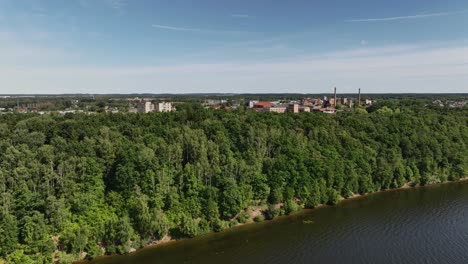 Vista-Aérea-La-Orilla-Del-Lago-Está-Cubierta-De-árboles,-Y-Detrás-De-Ellos-Se-Pueden-Ver-Los-Edificios-De-La-Ciudad