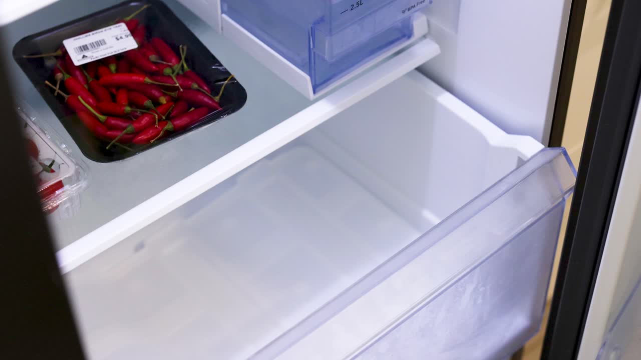 A person places fresh vegetables into a refrigerator, ensuring proper storage. Bright lighting highlights the organized kitchen environment