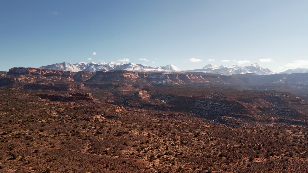 A high-flying drone shot over a remote dirt road cutting through the vast and unique desert land near Moab, Utah, with the snowy Rocky Mountains towering in the distance