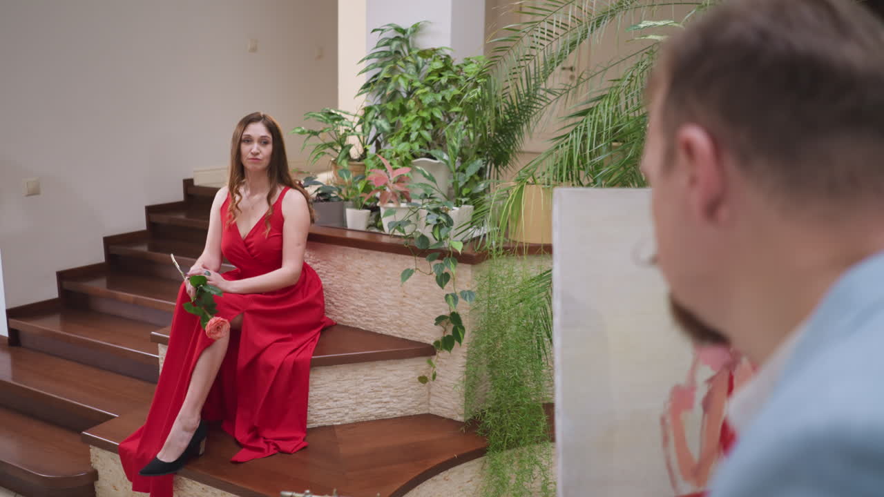 갤러리에서 예술가에게 포즈를 취하는 사려 깊은 여성, 빨간 슬리트 드레스를 입은 젊은 여성이 계단 에 앉아 있으며, 화가는 미술 스튜디오에서 그림을 그리고 있습니다.