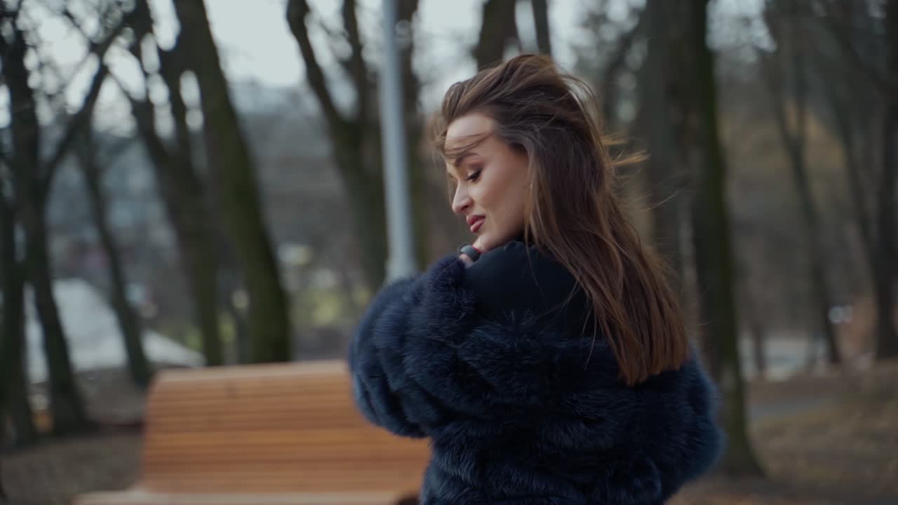 Attractive girl in the city park. Stylish woman in fur coat turning around and looking on camera outdoors. Slow motion.