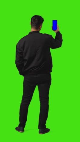 Vertical-Video-Full-Length-Shot-Of-Young-Man-Holding-Blue-Screen-Mobile-Phone-Standing-Against-Green-Screen-From-Behind