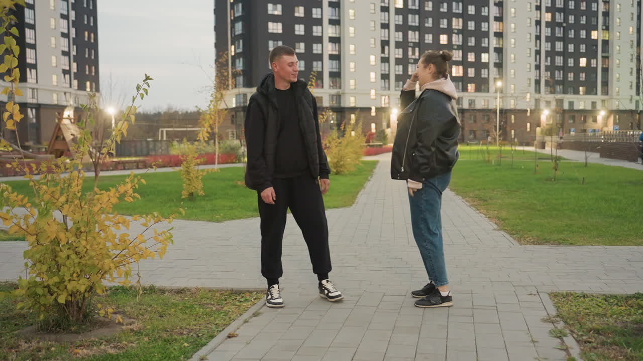 Playful woman enthusiastically performs martial arts move during evening walk in urban park as her amused friend stands nearby laughing and gesturing for her to stop so they can continue walking