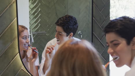 happy caucasian lesbian couple brushing teeth together and smiling in sunny bathroom