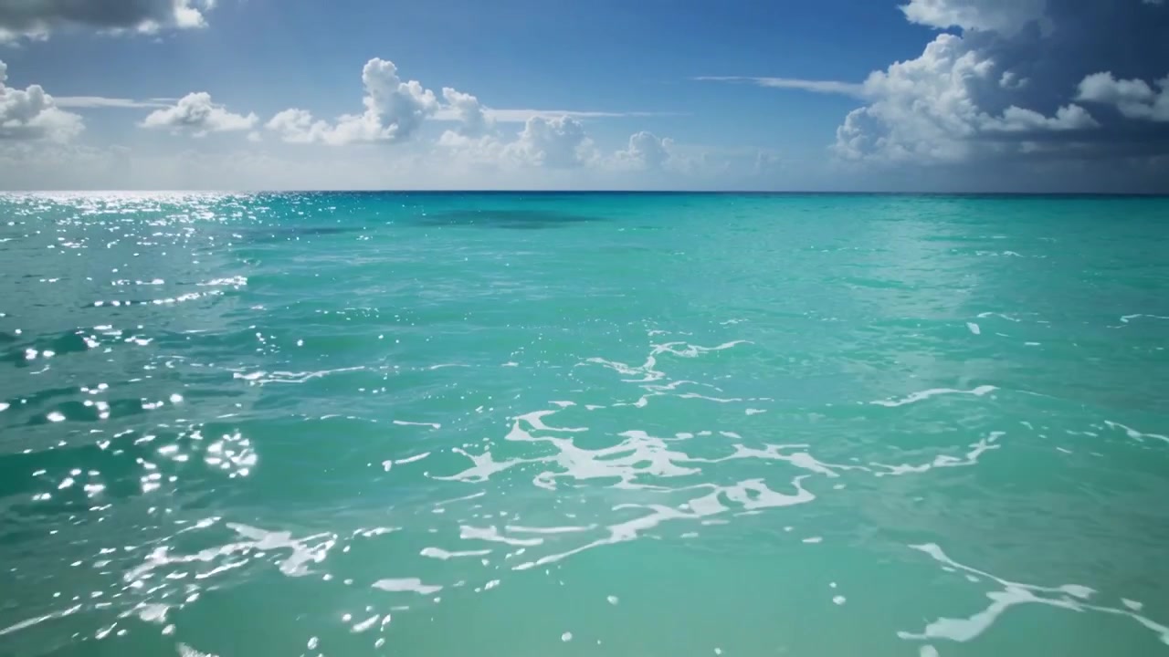 Türkisfarbene Meeresoberfläche rollt im Wind, glitzert, treibender Schaum bewegt sich zum Ufer, Sand wird sichtbar.