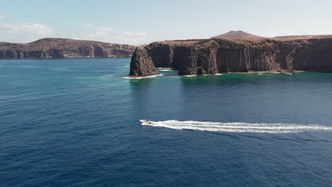 Roque-Partido,-Gran-Canaria:-Vista-Aérea-Con-Seguimiento-Lateral-Sobre-El-Barco-Y-El-Majestuoso-Roque-Partido-Al-Fondo-En-Un-Día-Soleado