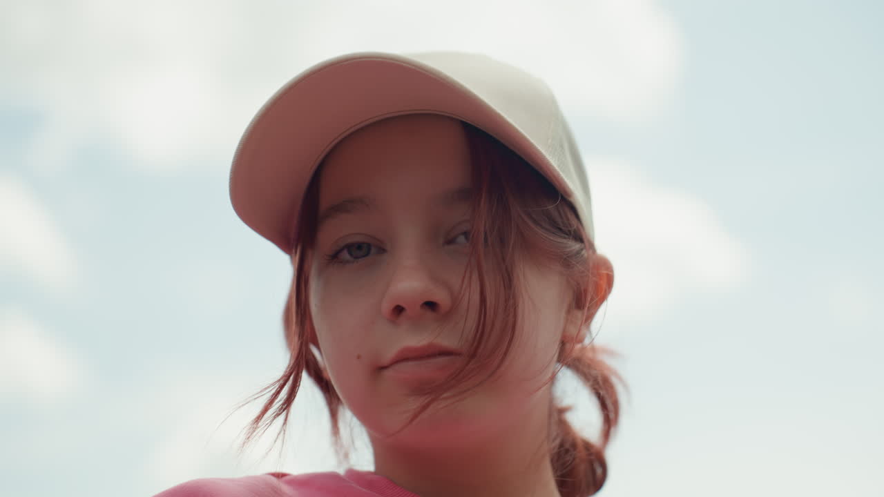 White Teenage Girl Wearing Cap Pensive, Low Angle Closeup Against Blue Sky And Modern Building, Soft Cinematic Tones, Red Hair Strands Framing Face, Subtle Sunlight, Introspective Mood And Urban