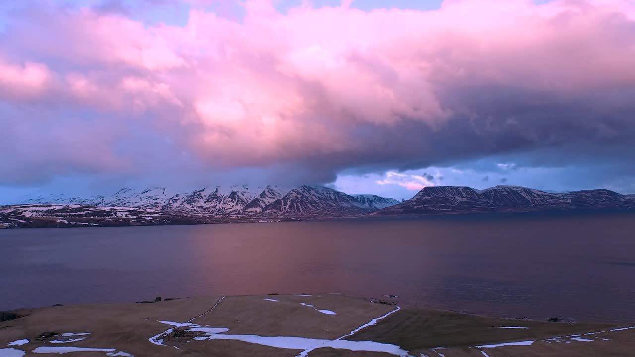 An aerial shot of pink clouds float over a lake with snowy mountains in the background in Iceland
