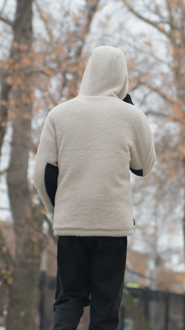 겨울 재을 입은 젊은 성인이 고 전화로 이야기하는 뒷면, 건조한 나무, 건물 및 불분명한 구조의 흐릿한 배경, 추운 날 도시 환경