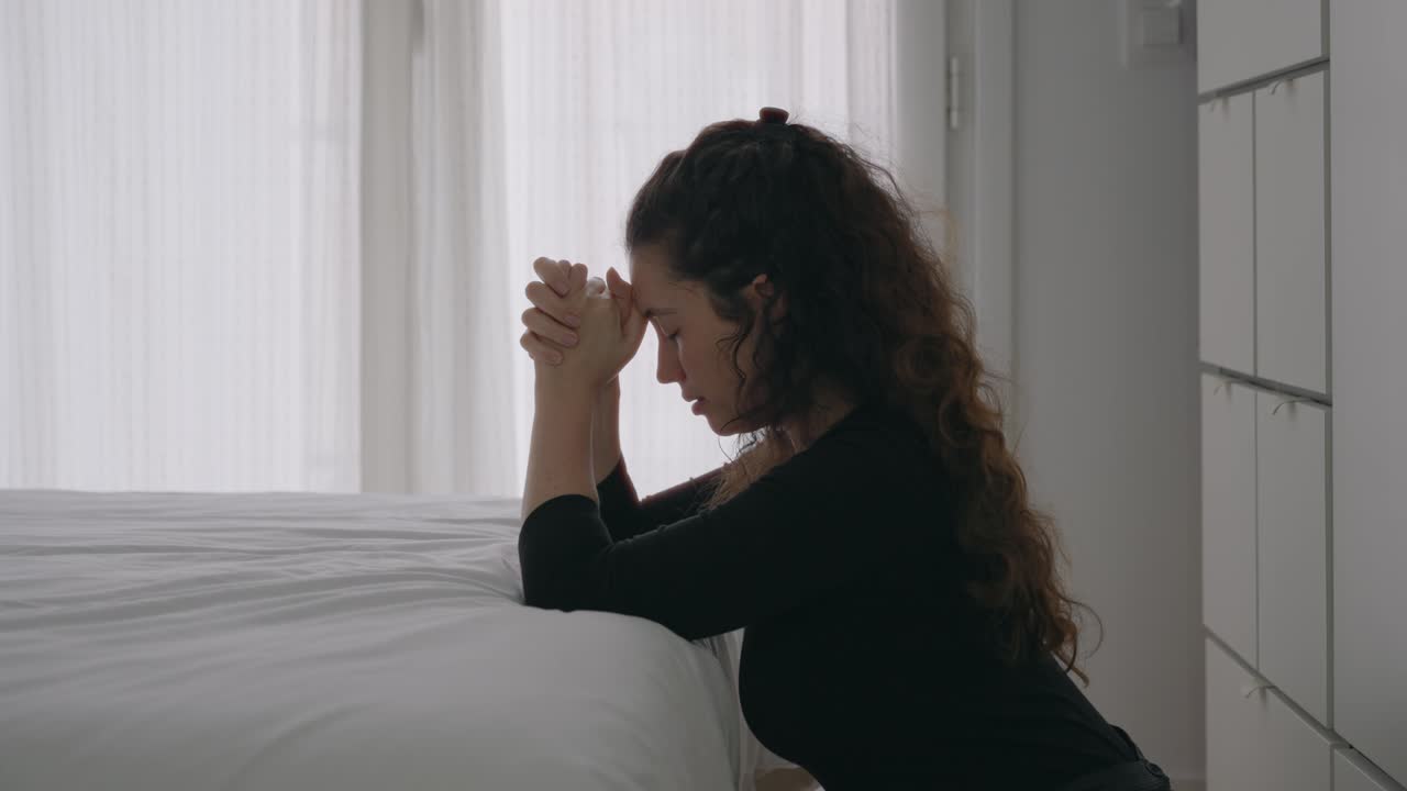 Religious female believer is praying while kneeling by the bedside in her bedroom during daytime. Sun light shining from outside through window curtains. Bright light. Her eyes closed
