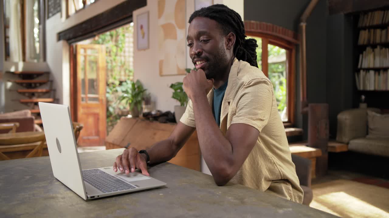 African American man sitting at home starting work on silver laptop, typing, pausing, smiling