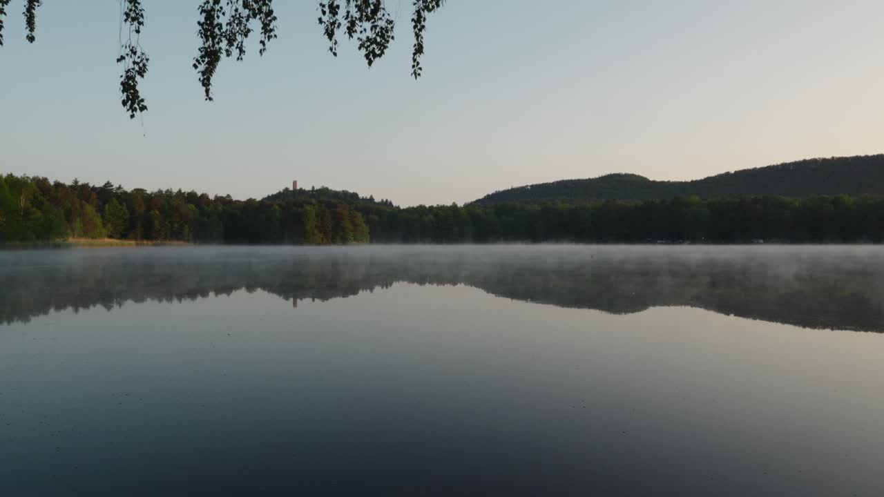 부드러운 안개가 천천히  ⁇ 은 호수를 가로질러 움직여 다른 쪽의 숲 언덕을 반영합니다.