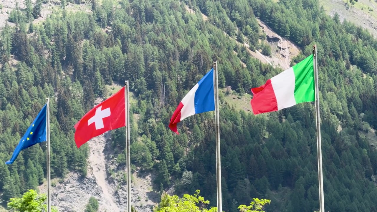 Bandiere dell'Unione Europea, svizzera, francese e italiana sventolano su uno sfondo montano alpino boscoso.