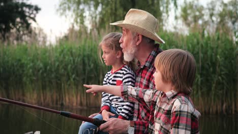 close-up view of a boy showing something to his grandfather and his little sister while they are fishing sitting on the lake pier