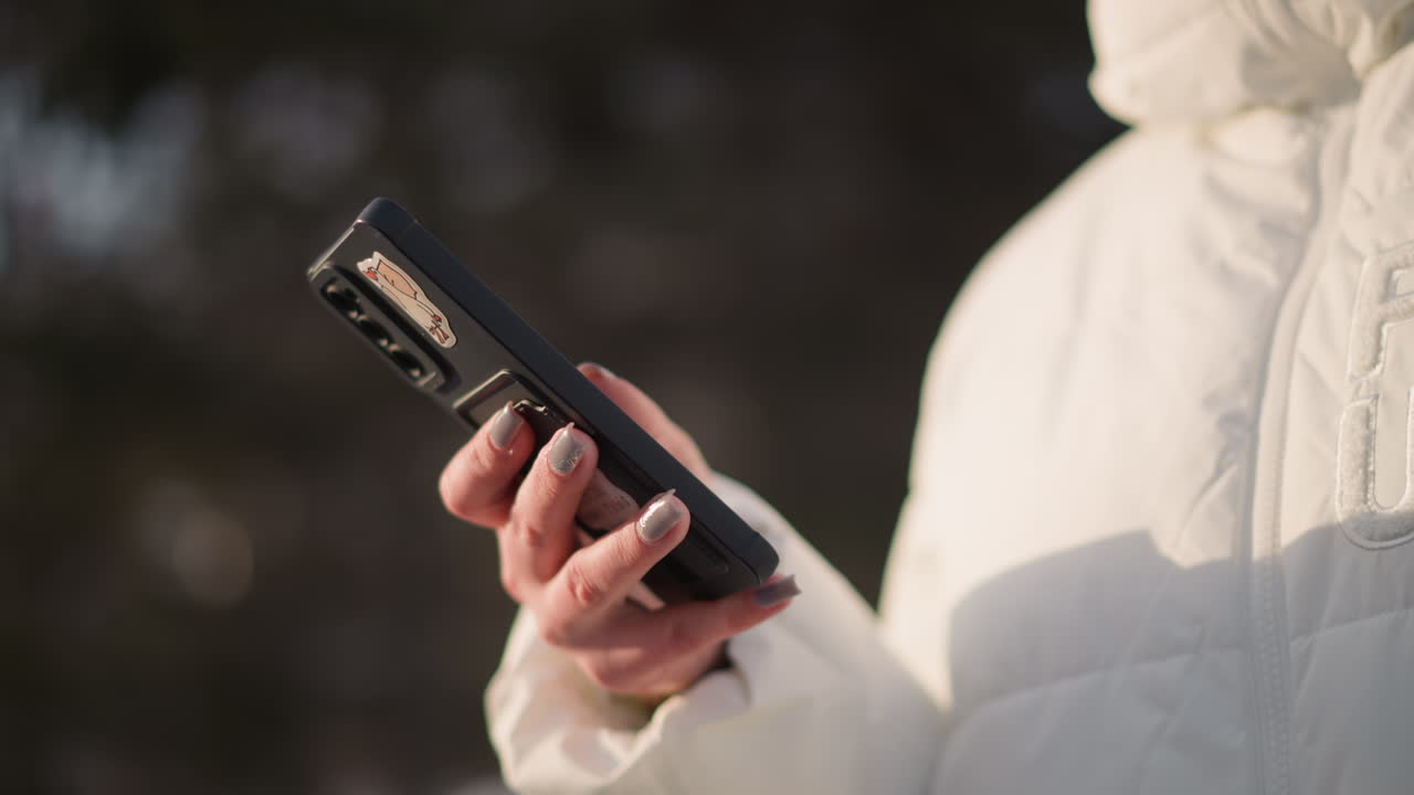 Content creator scrolling smartphone screen with manicured nails against white winter jacket in snowy outdoor park setting, capturing tech lifestyle vibe under soft daylight glow