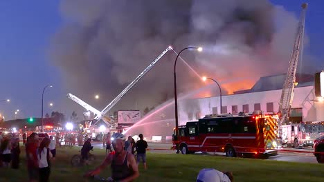 firefighters battling a large fire, with smoke billowing into the sky as crowd of onlookers watch at dusk