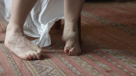 a white woman seated on a wooden chair, she's bare feet and playing with her toes on a red carpet